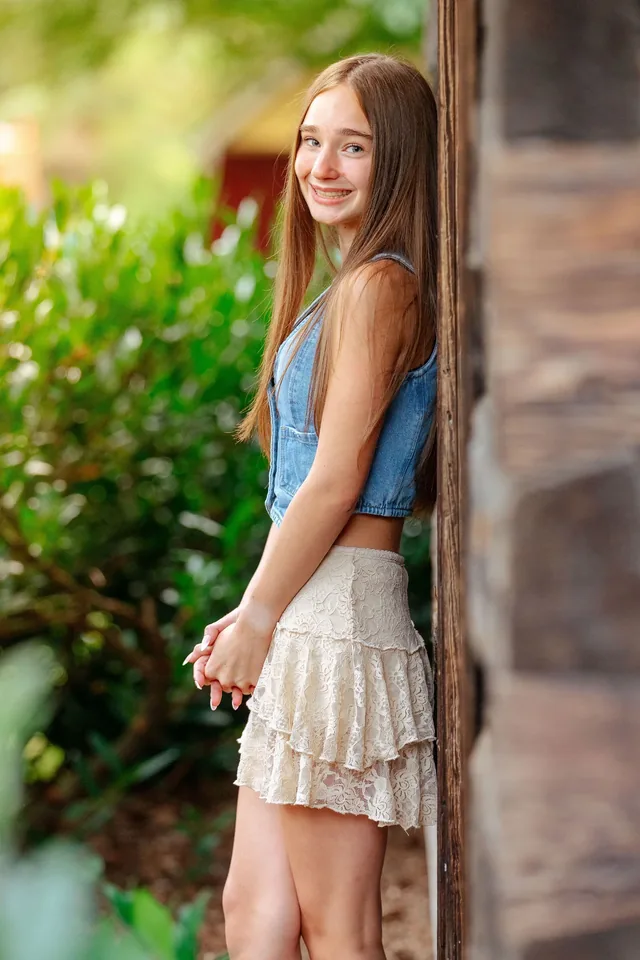 Senior in a denim vest and lace skirt smiling beside a log cabin wall in warm evening light
