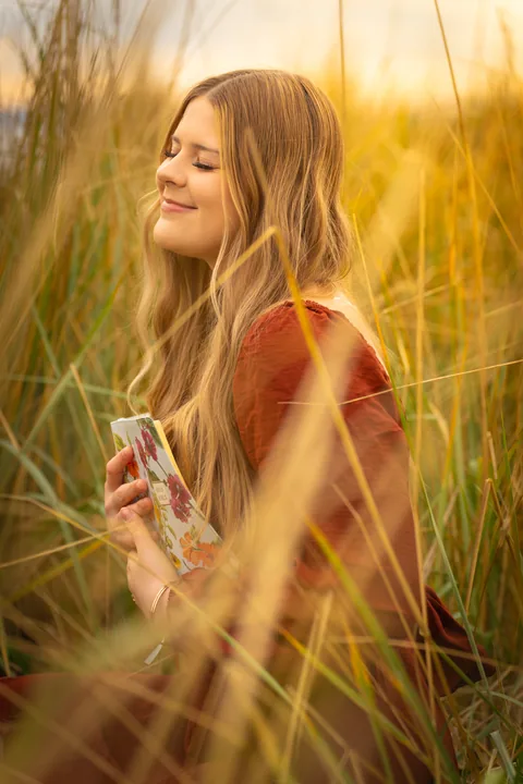 Senior in a rust dress holding a book in tall golden grass at sunset