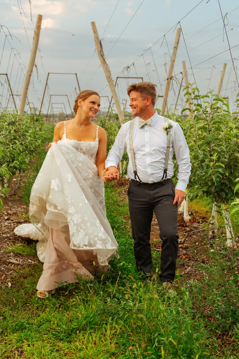 Bride holding her dress as the couple walks the orchard rows